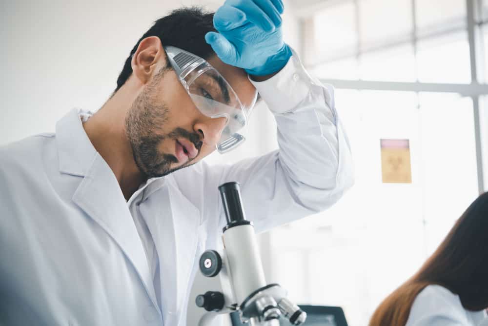 Male scientist wiping arm across forehead while standing by microscope