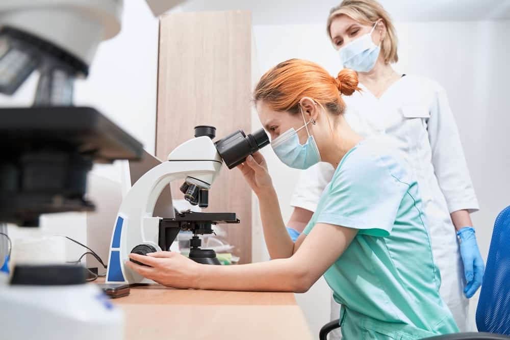 Woman in medical scrubs and mask using microscope while woman in lab coat watches