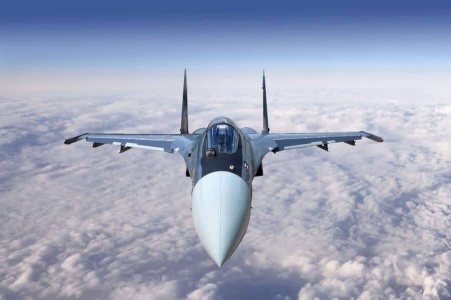 Military jet soaring above white clouds in a clear blue sky.